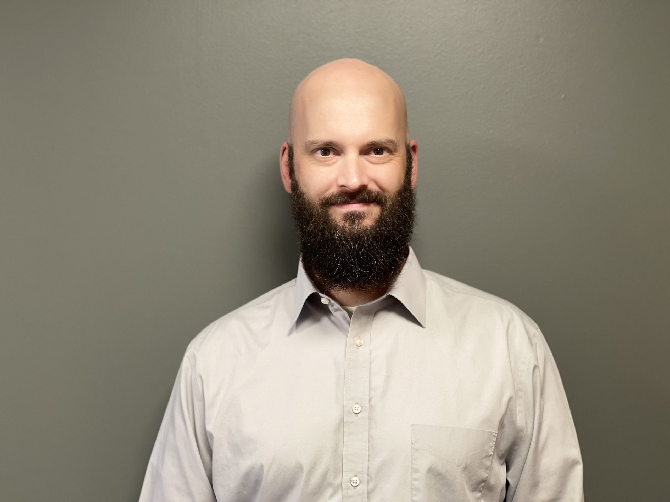 a white bald man with a dark black beard stands in front of a grey wall. He is wearing a kind expression and a light grey button up.
