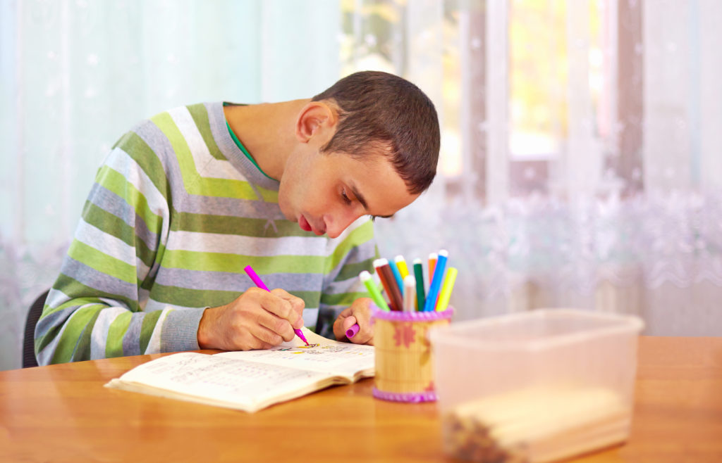 A man with developmental disabilities uses a marker to fill in his journal. He has light skin and very short brown hair. He is wearing a green and grey striped shirt, sitting at a table with a jar of markers and bucket of pencils on it.