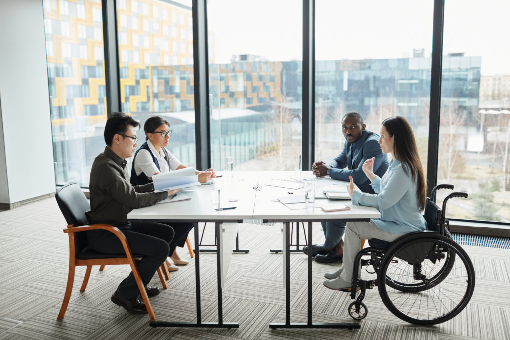 Wide angle view at diverse business team discussing project at table in modern office with focus on young woman in wheelchair sharing ideas in foreground.