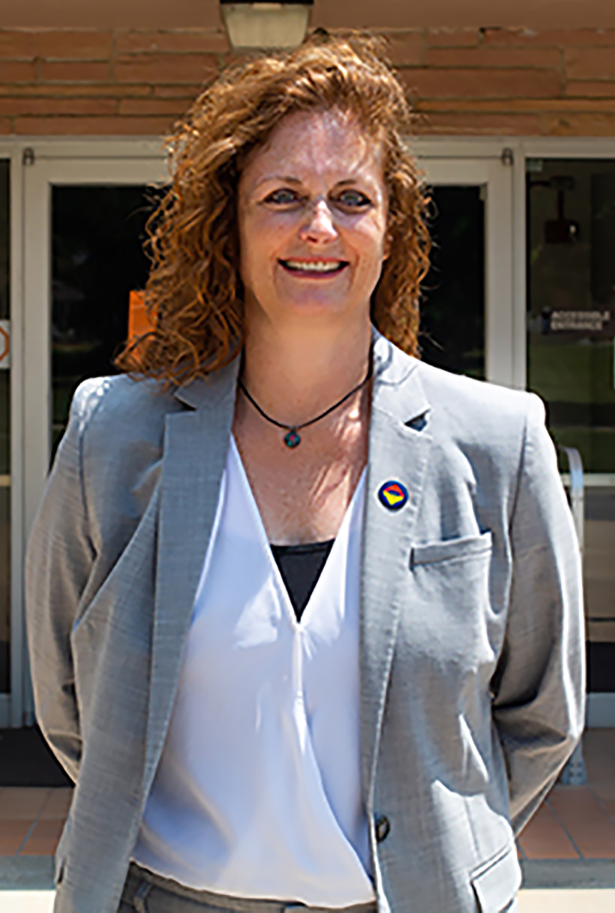 White woman with red curly hair stands outside of a brick building in a grey suit jacket and white blouse. She is seen from the waist up and is smiling at the camera with her hands behind her back