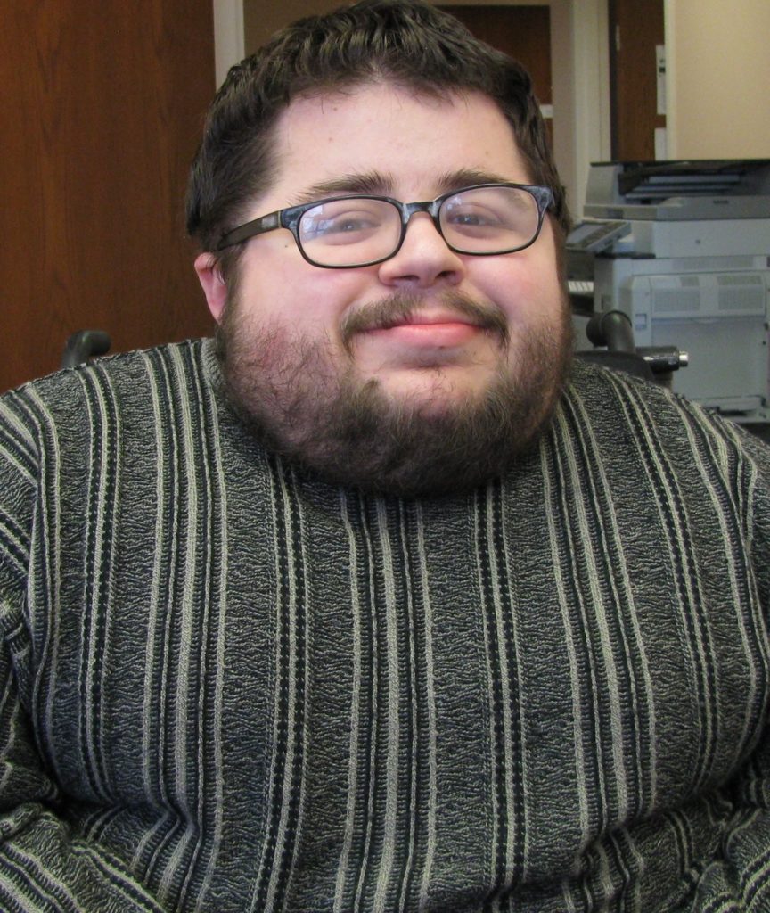Emmanuel sits in front of their office furniture. They are wearing a green vertical striped sweater and plastic rectangular glasses. They are white with dark black hair and a beard.