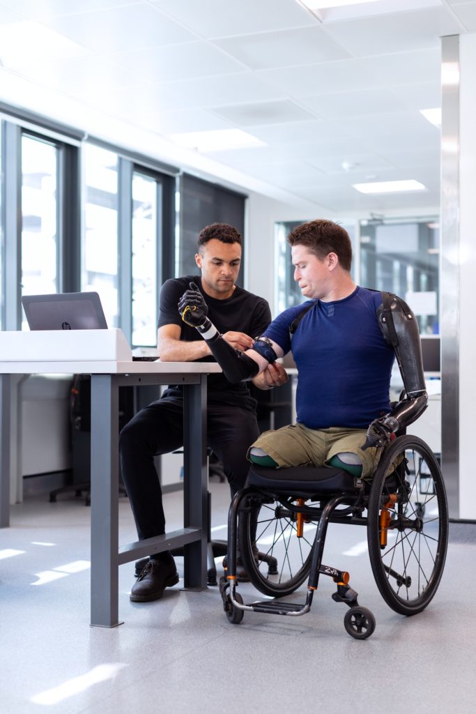 black man is fitting a with man with the technology required to move an arm prosthetic. The white man is quadriplegic and is wearing a dark blue cotton t-shirt and camel colored cargo shorts. He is sitting in his wheelchair. The man fitting the technology wears all black.