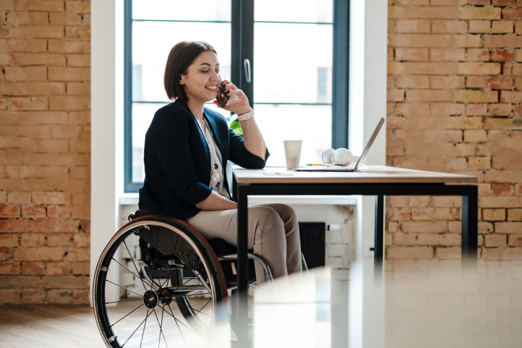 Young disabled business woman in wheelchair working at office desk and with laptop, talking on mobile phone 