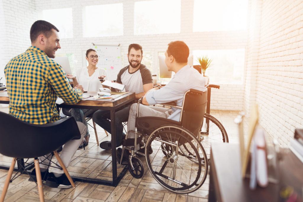 Office workers and man in a wheelchair are making a conversation in bright office. They are showing a teamwork.