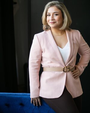 dior vargas stands with one hand on her hip and one to her side. she is a light skinned latina woman with shoulder length blonde hair and she is wearing a pink blazer and white v-neck shirt. She smiles with her head turned toward the camera with a trusting and silly soft smile.