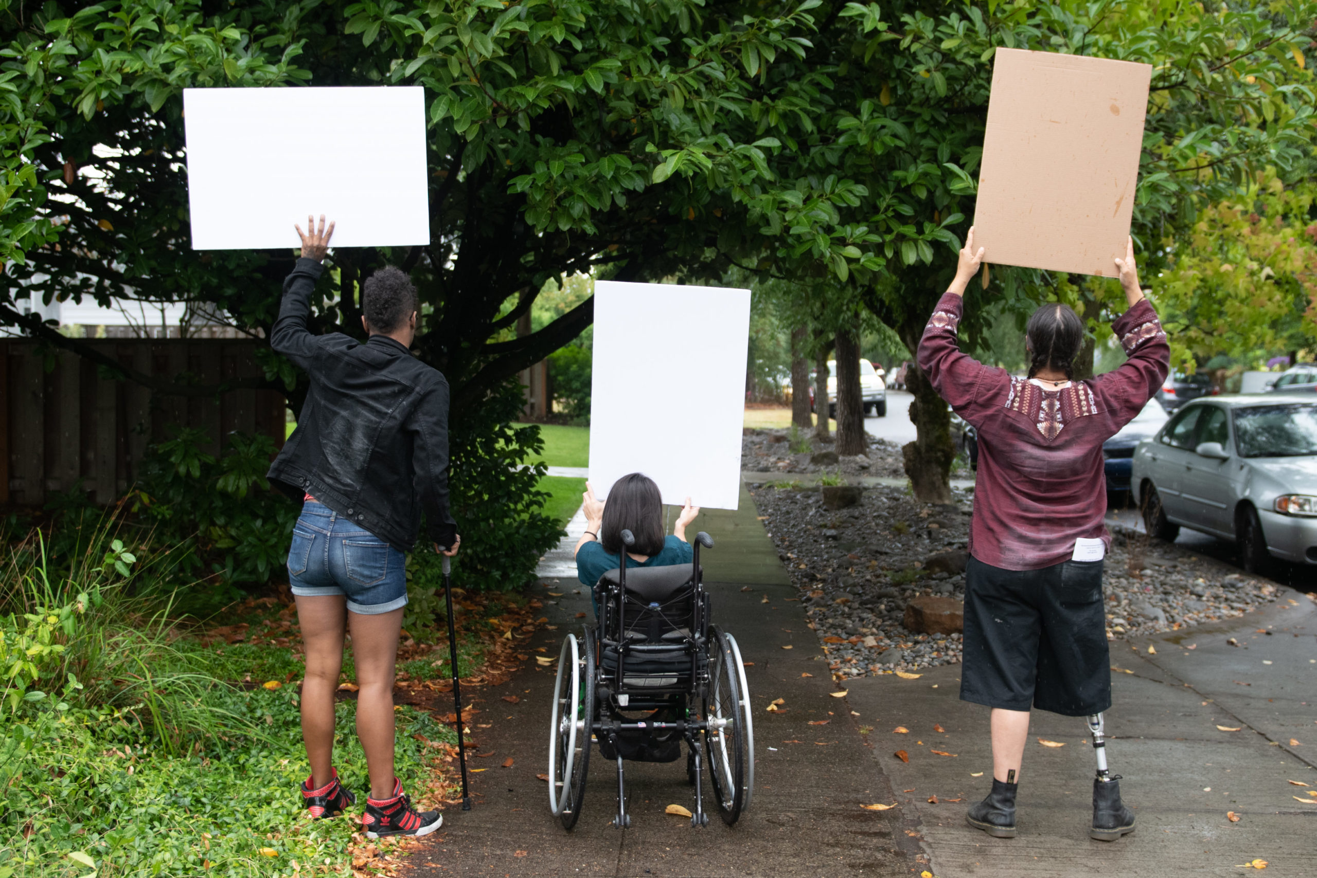 Three disabled people of color (a Black non-binary person with a cane, a South Asian person in a wheelchair, and an Indigenous Two-Spirit person with a prosthetic leg) block a neighborhood street while holding up cardboard signs. The photo is shot from behind everyone.