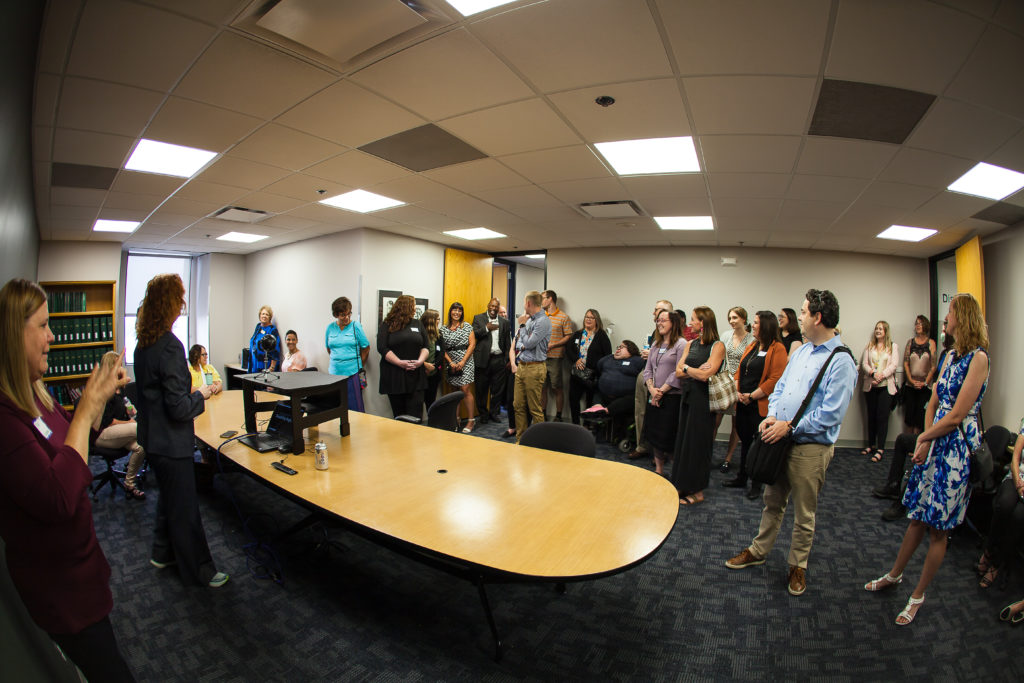 A large group of people congregate in a conference room all facing a speaker. The speakers back is to the camera. 