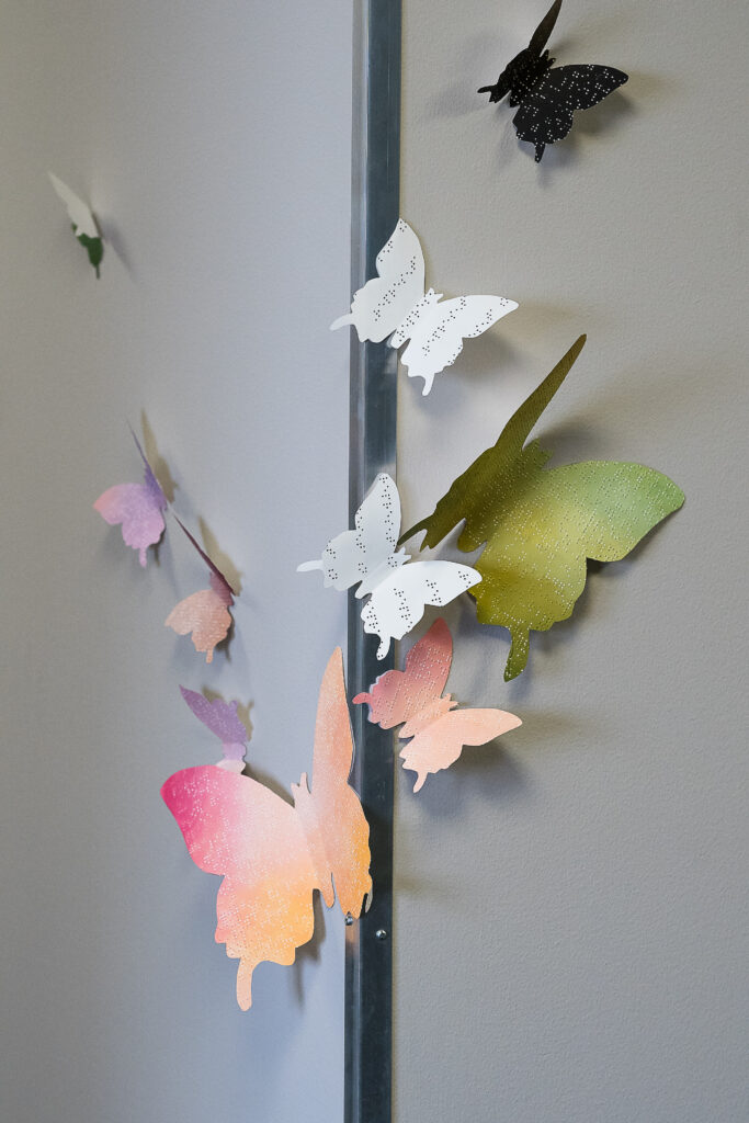 cutout butterflies with braille and painted colors on top of them spread out from the corner of a wall all flying away from the corner.