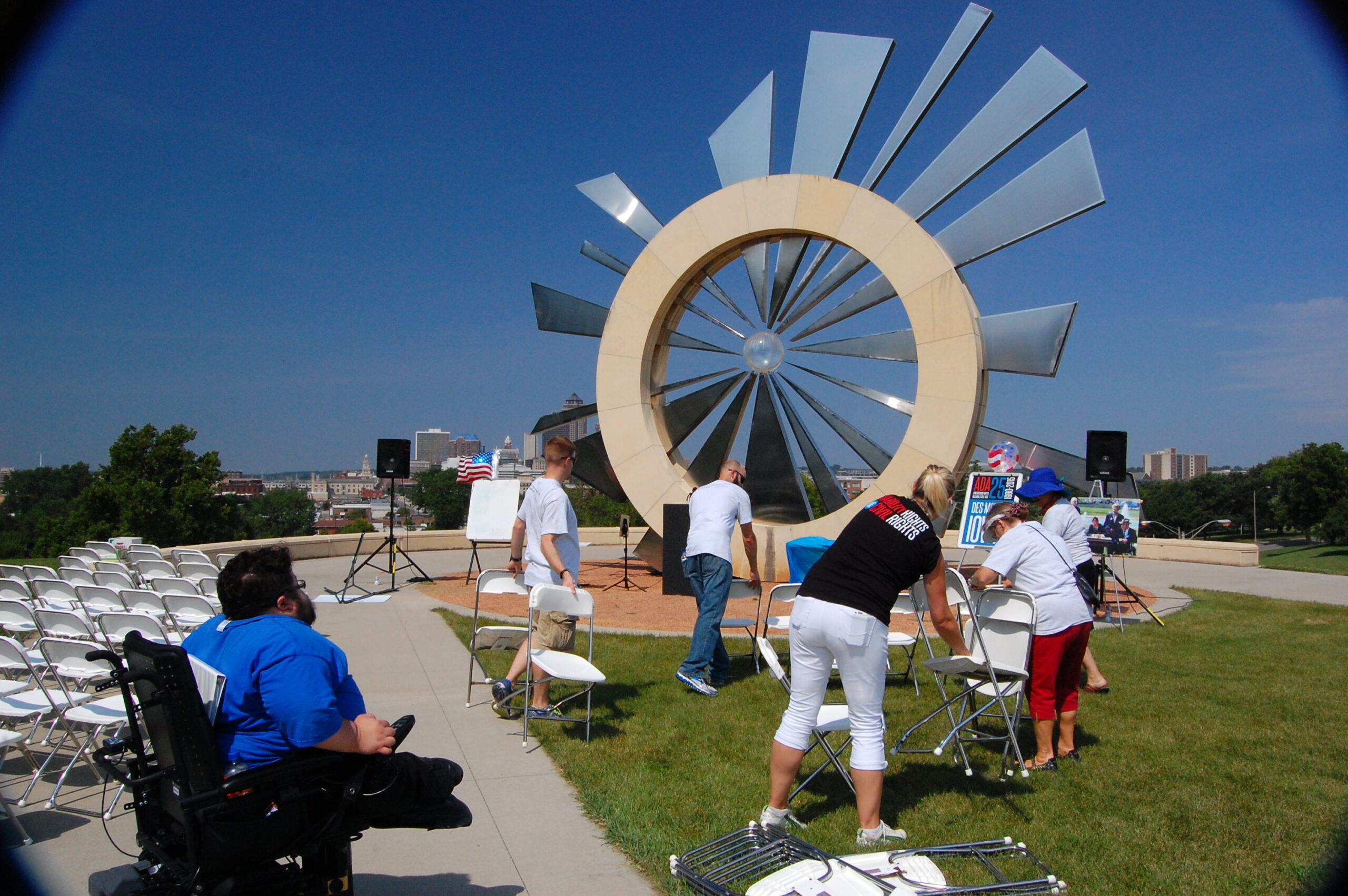 people with disabilities are standing and sitting around a lawn setting up for an event in front of a large round and spoked art sculpture.