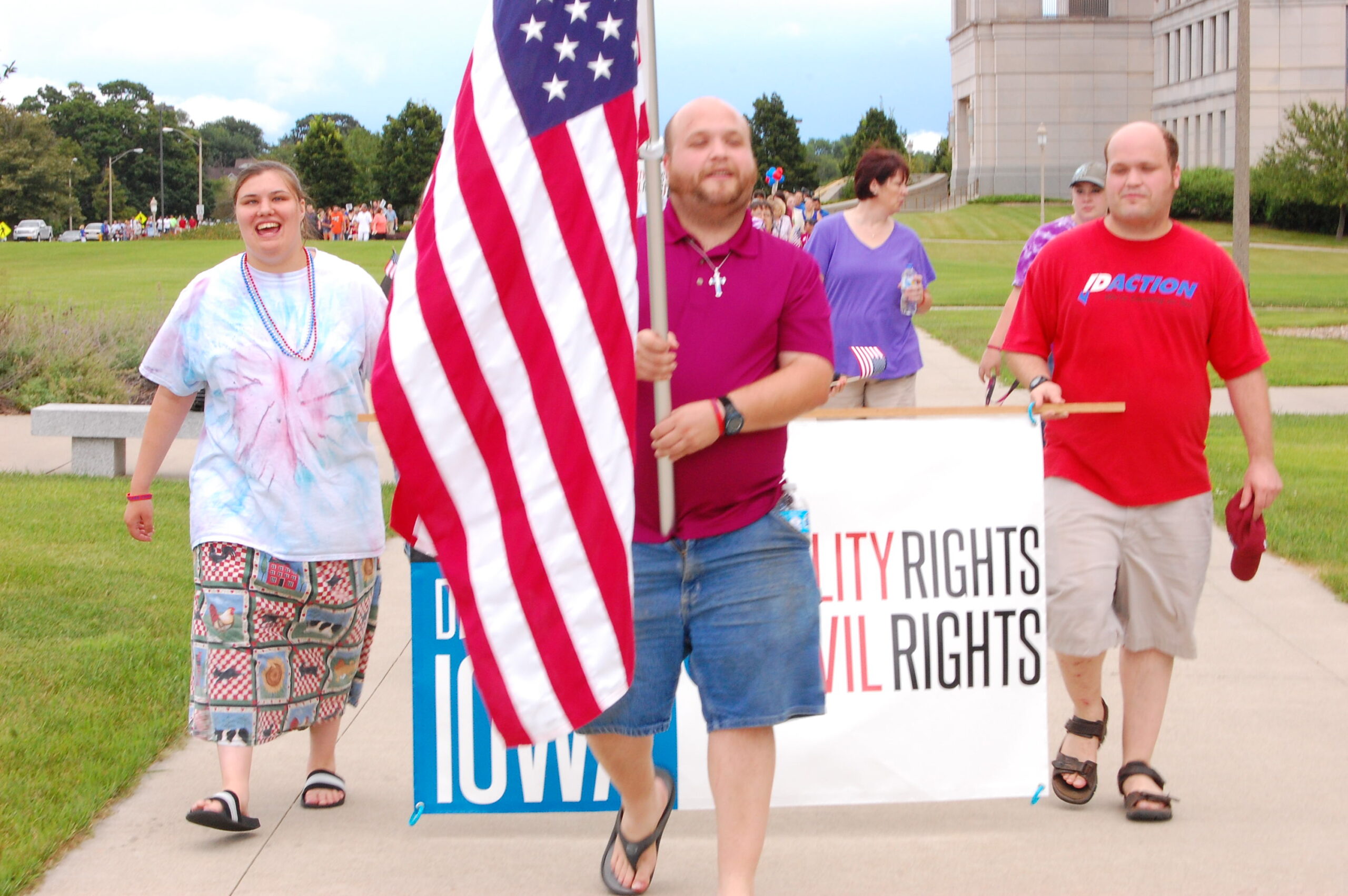 Image of a man walking with the American flag in his hands. He is in front of a group of people also walking in the same path, holding a "disability rights, civil rights" poster.