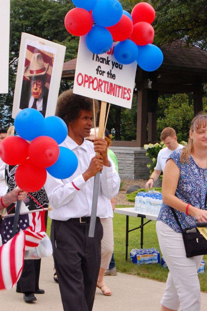 Black man stands holding a sign that reads "thank you for the opportunities" with red and blue balloons filling the screen.