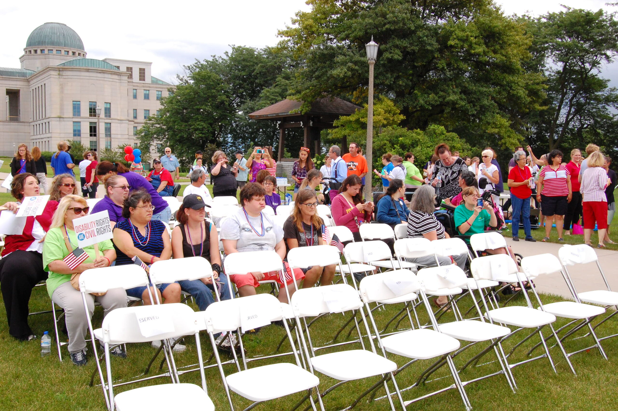 several rows of folding chairs are arranged in a yard. There are people standing around and sitting in the chairs.