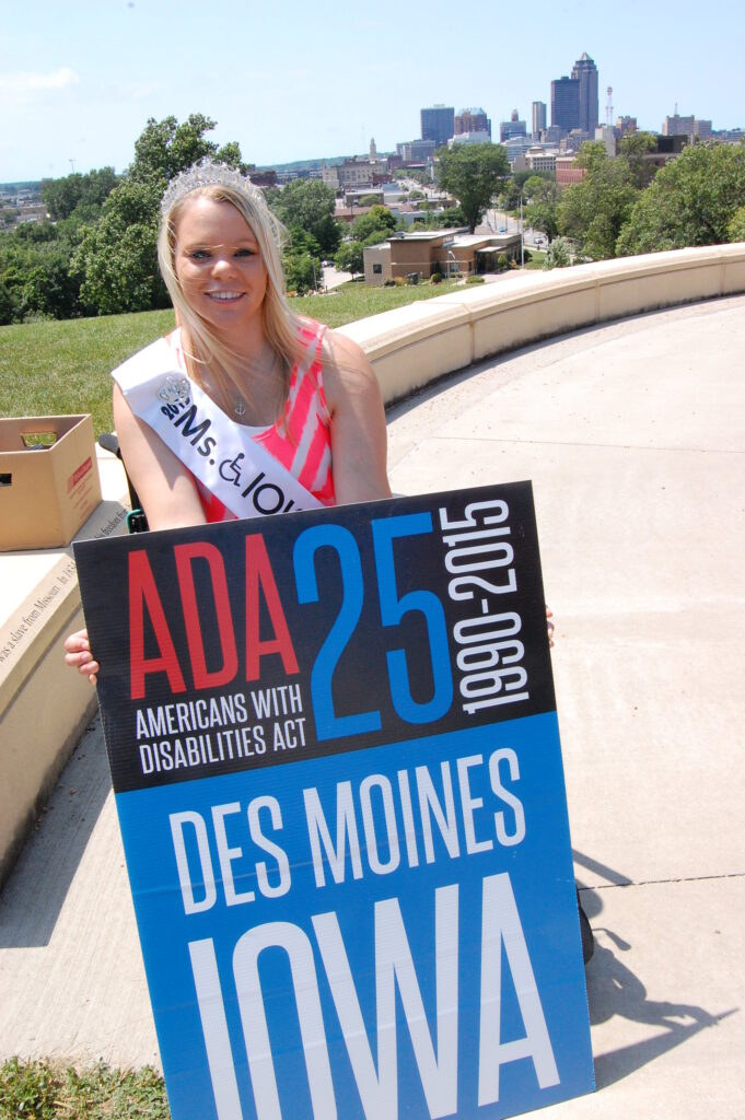 Ms. Wheelchair Iowa sits holding a sign that reads "ADA 25 1990-2015, Des Moines Iowa" She is wearing a dress and her tiara. 