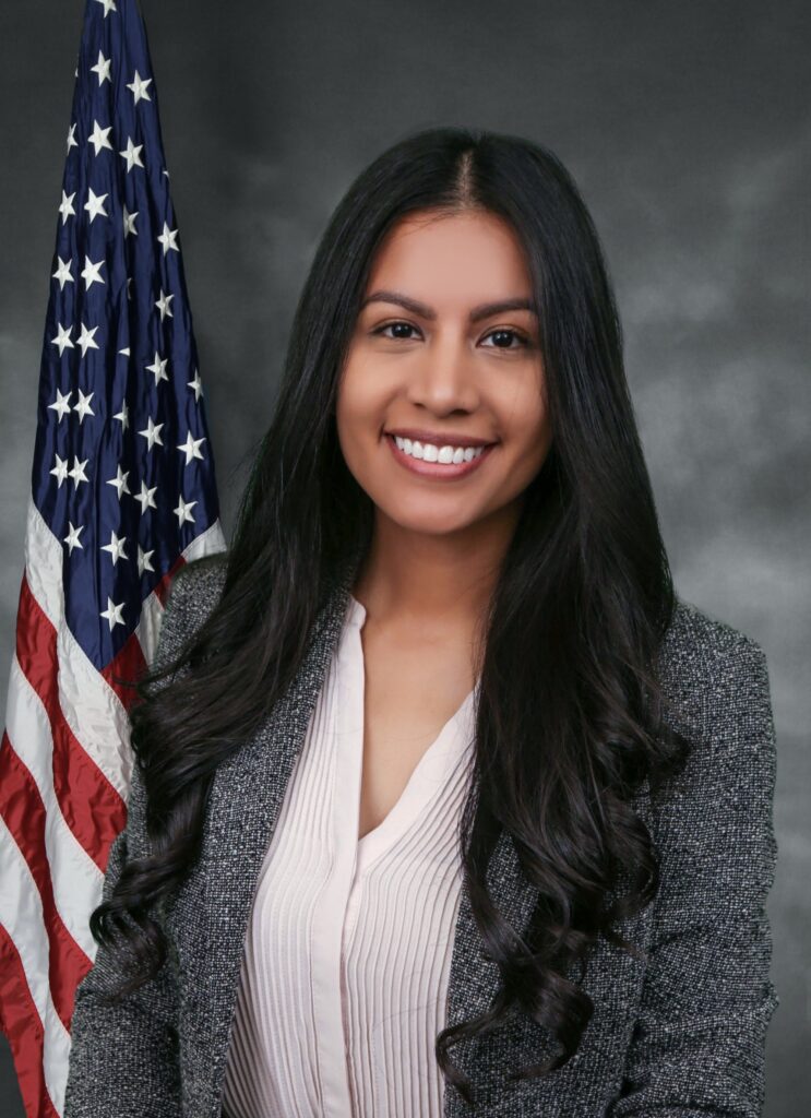 Gloria is in front of a gray background with an American flag hung behind her. She has long black hair that is curled all the way to the bottom of the photo. She is smiling at the kindly at the camera.