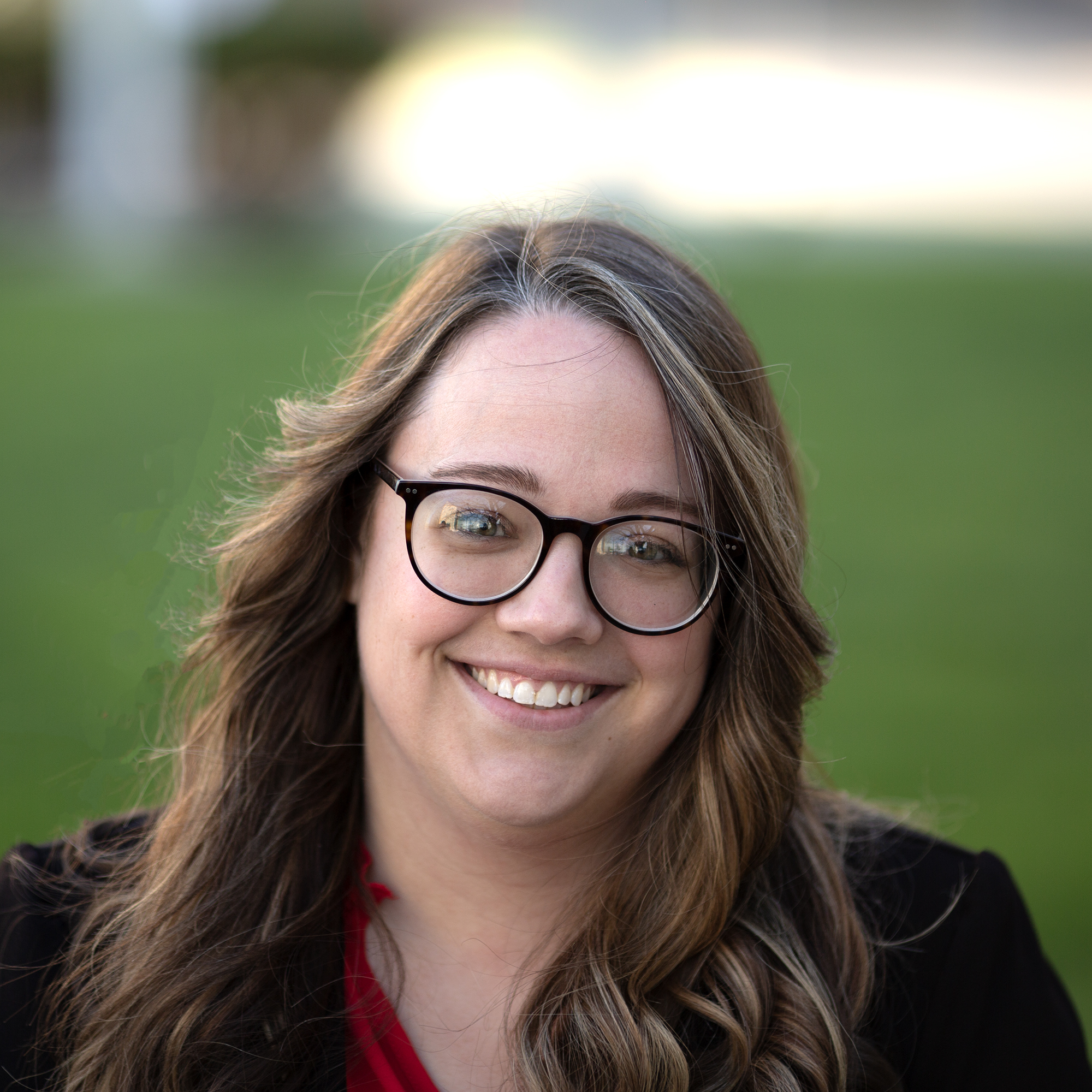 Steph is a white woman in her late 20s with long brown curled hair. She has on round plastic glasses and a blazer. She is smiling in front of a blurred background.