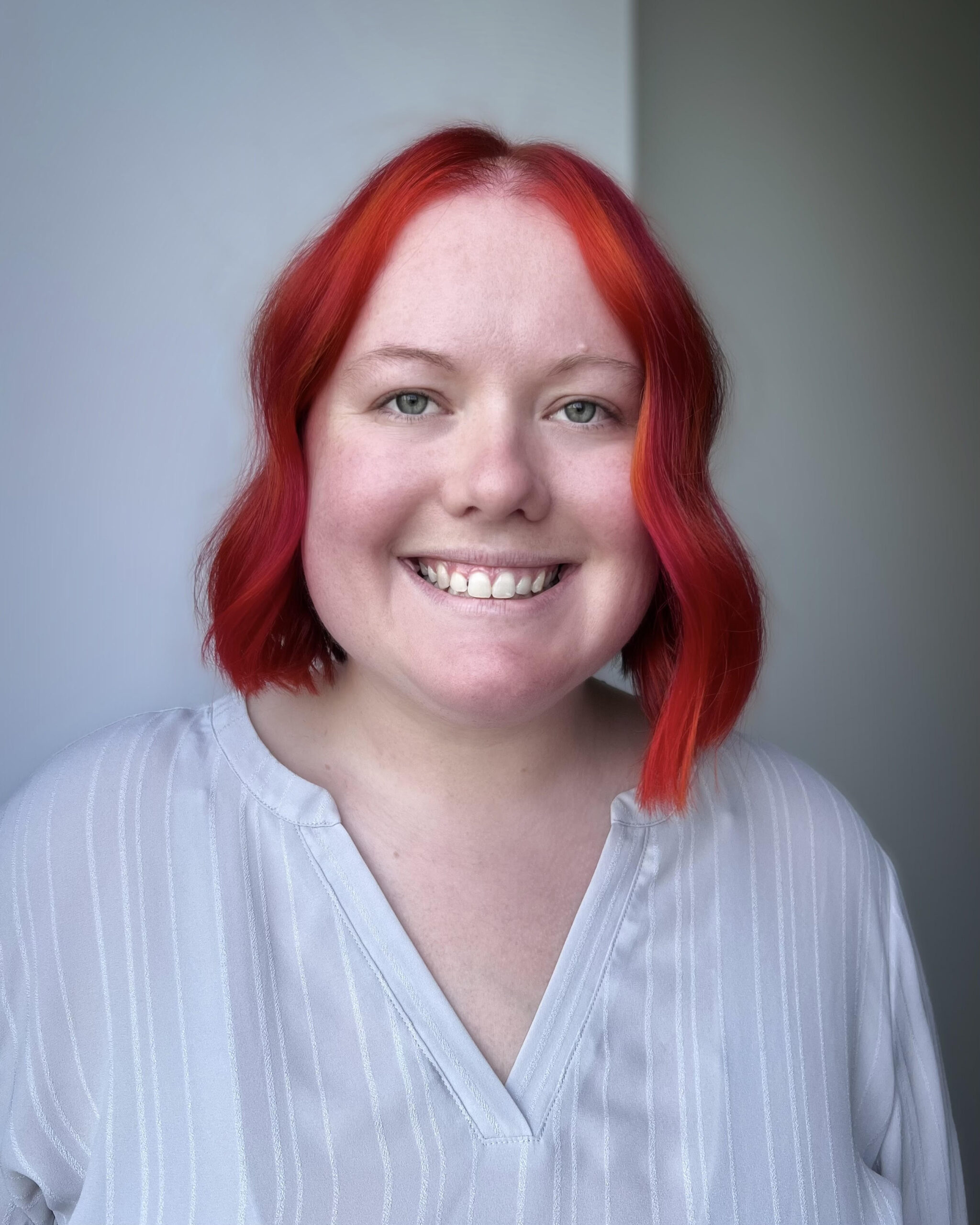 Jenny is a white woman in her late twenties with bright red hair that is cut right above her shoulders. It is curled slightly. She has bright eyes and a bright smile. She is pictured in front of a light white wall and is wearing a light white shirt.