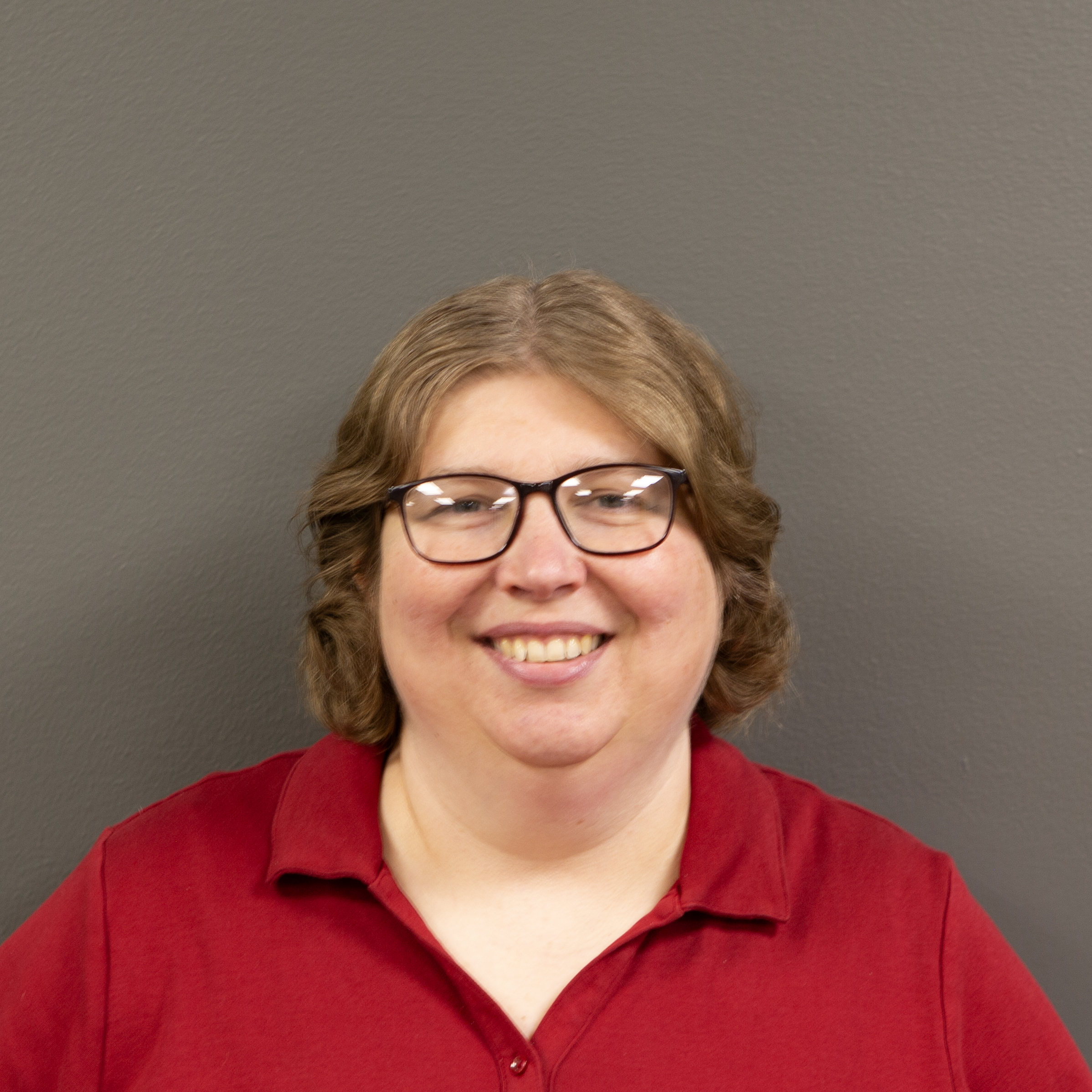 Tammy is a middle aged white woman with curly light brown hair that is cut to the middle of her neck. It is parted in the middle. She wears rectangular glasses that are dark and plastic. She is smiling kindly and wearing a bright red polo shirt while standing in front of a solid gray wall.