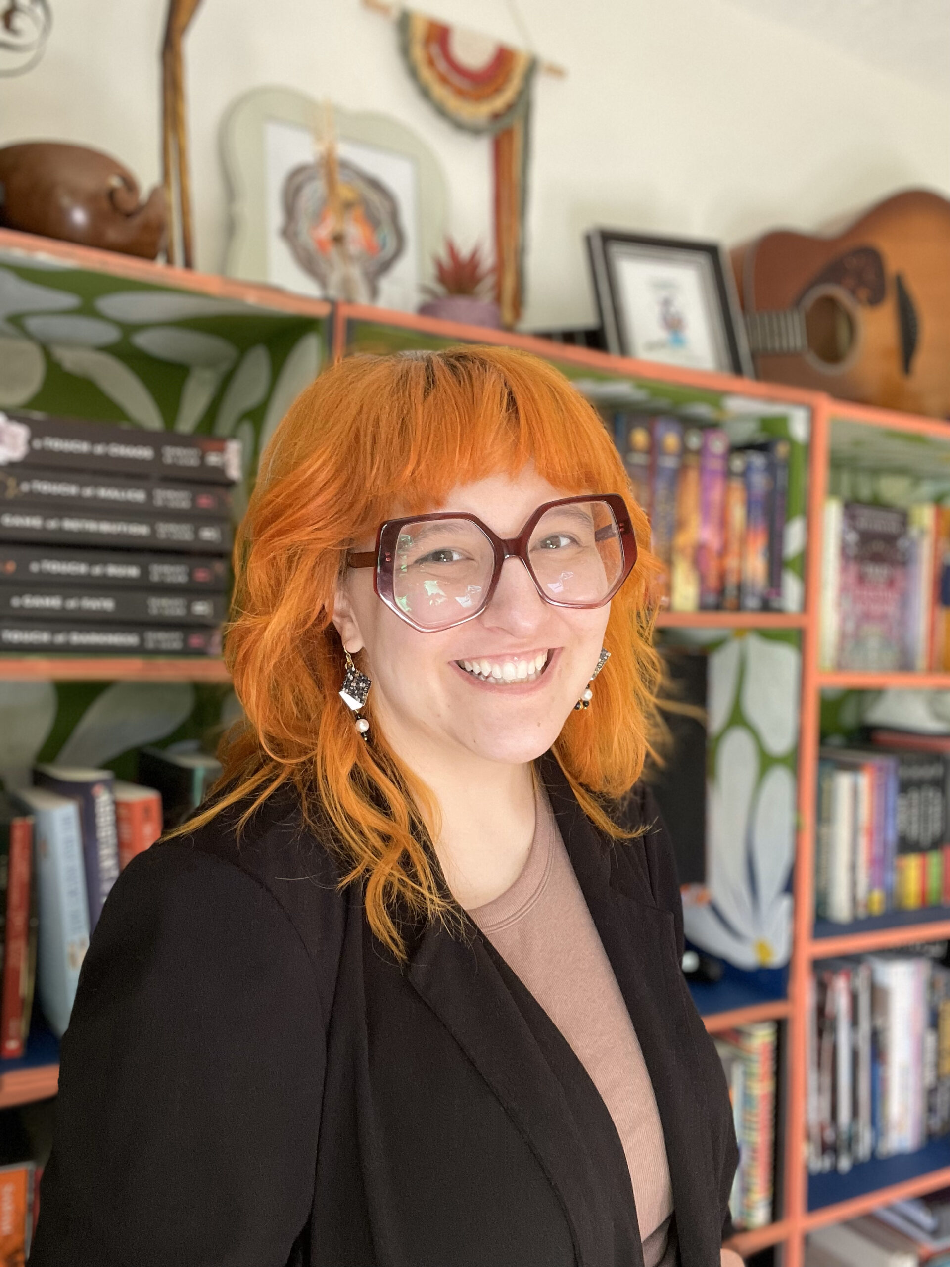 light skinned young woman stands with a wide smile and raised eyebrows in front of colorful daisy patterned bookshelves. photos, plants, crochet, and a guitar are on top of the shelves. She has bright orange hair that is curled past her shoulders with blunt bangs above hexagonal burgundy plastic glasses. She wears book earrings, a black blazer and a tan shirt.