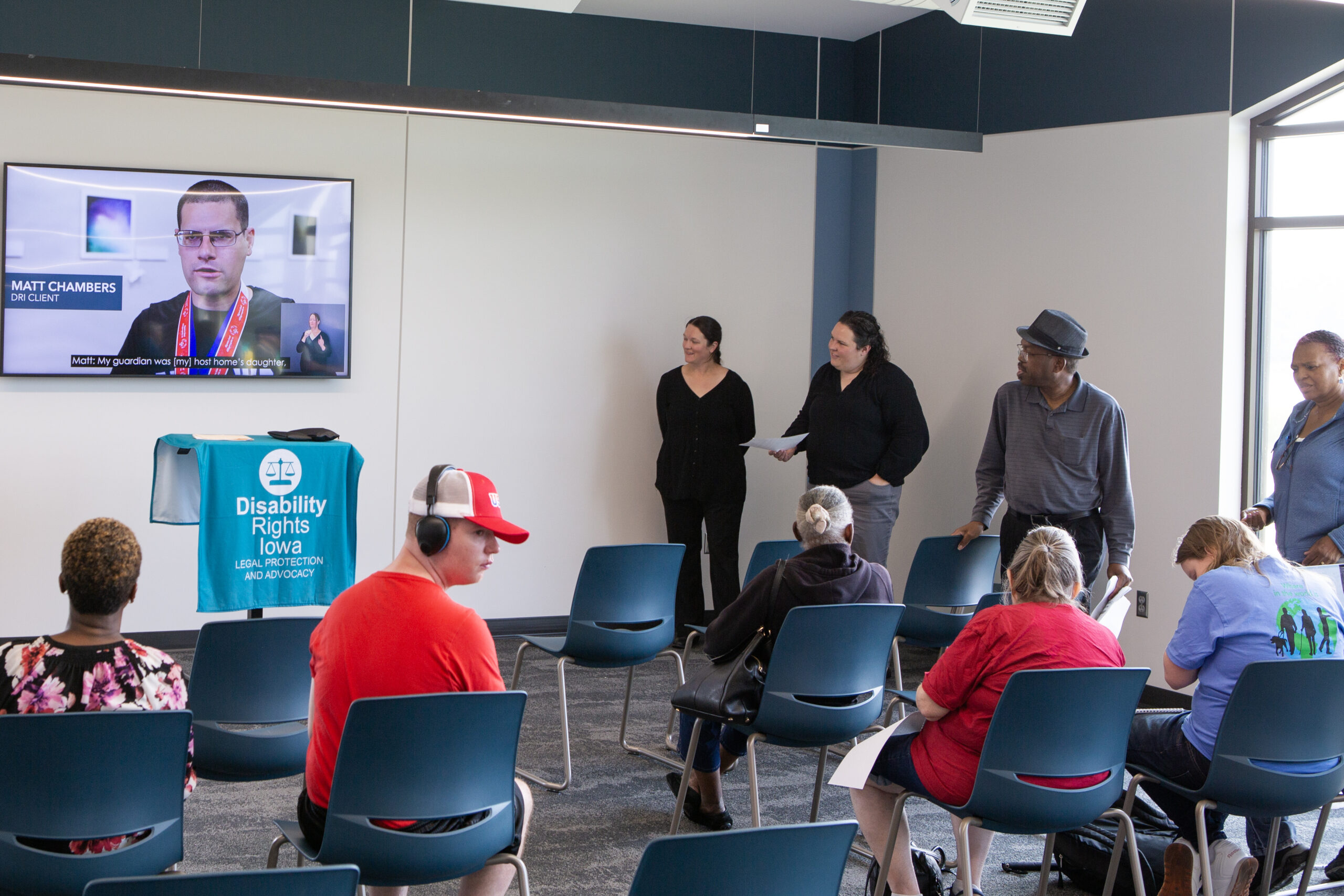 group of individuals sitting facing a tv screen with an individual giving an interview showing. asl interpreters stand to the side of the tv screen interpreting the tv interview.