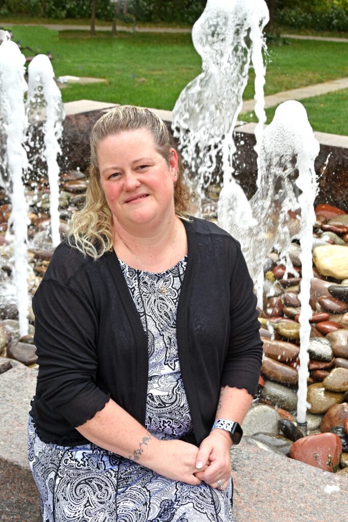 a middle aged white woman with blonde curly shoulder length hair sits in front of a water fountain. She has a soft smile on her face and is wearing a black and white curly patterned dress and black blazer.