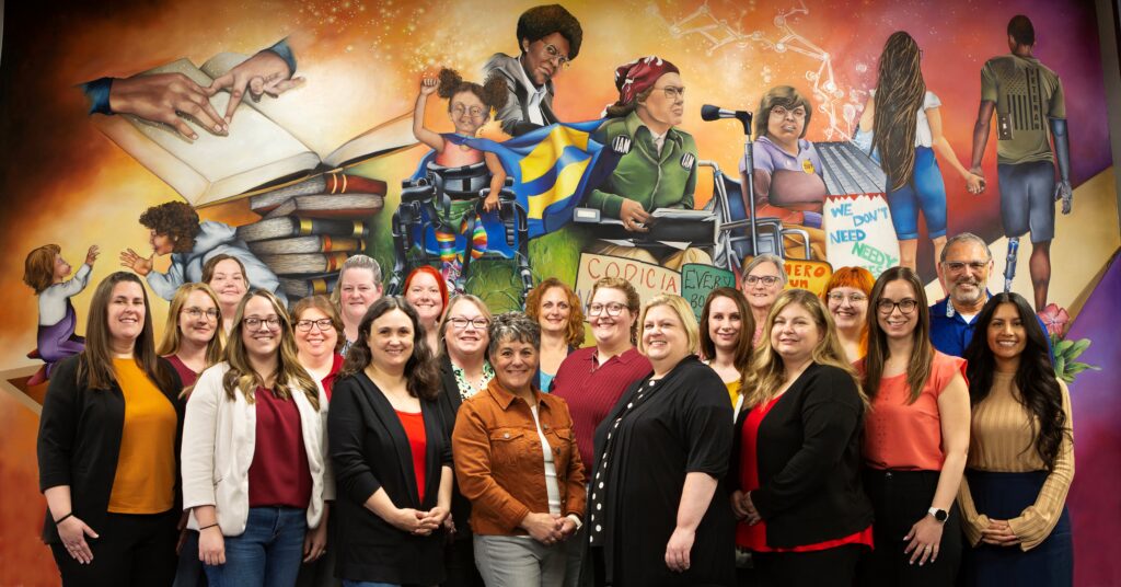 group photo of office staff standing and sitting in front of our Jill Wells disability inclusion mural.
