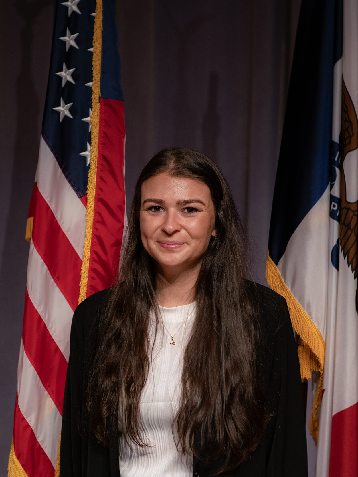 white woman with long brown hair split down the middle stands in front of an American flag, the Iowa flag and a curtained off wall.