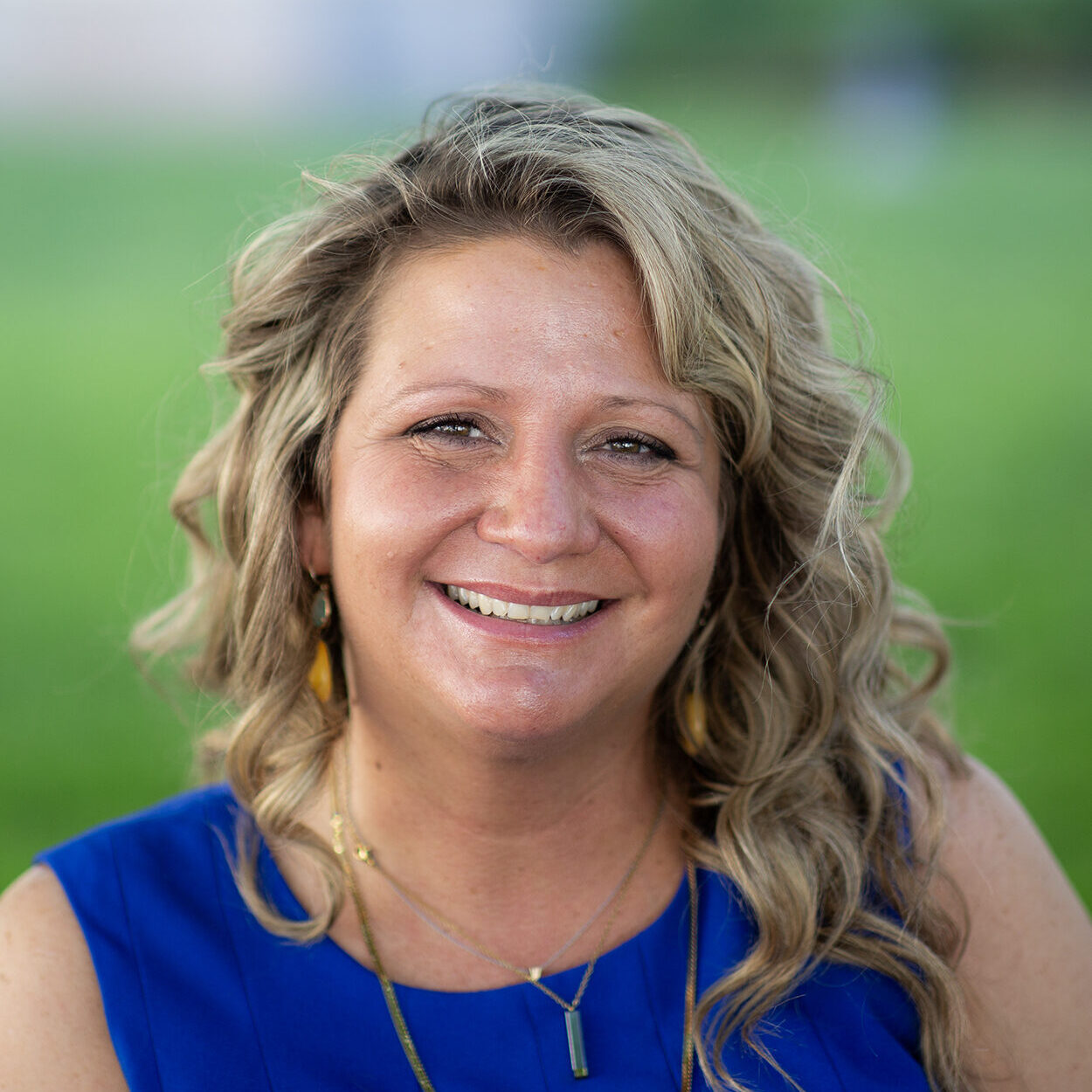 white woman with bouncy curled blonde hair stands in front of a blurred grassy background. She's smiling with her eyes crinkled and is wearing a blue sleeveless dress and a gold necklace.