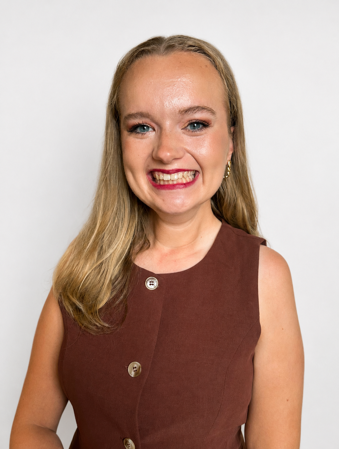 white woman in her mid twenties with shoulder length blonde hair smiles wide at the camera while standing in front of a white wall.