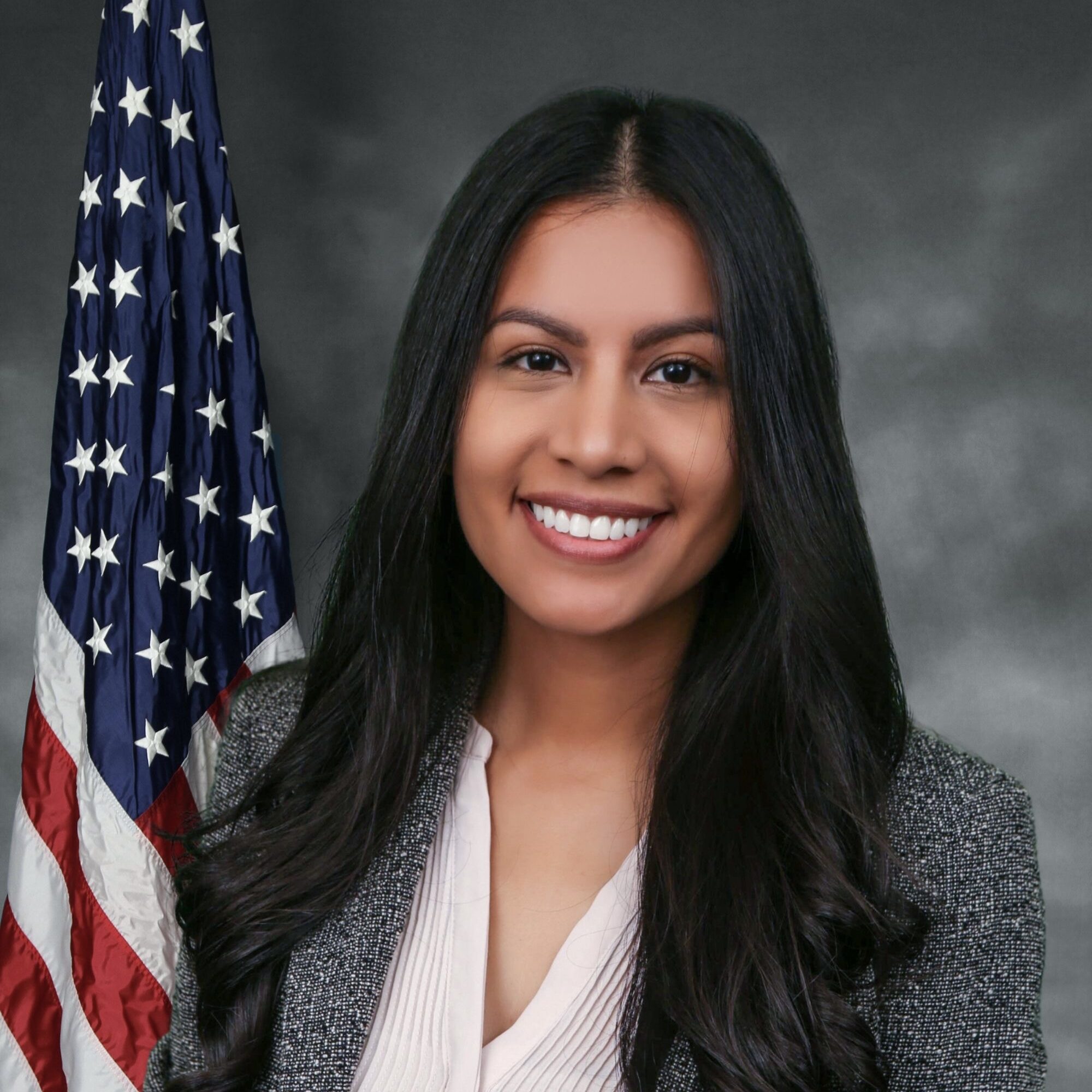 Gloria is in front of a gray background with an American flag hung behind her. She has long black hair that is curled all the way to the bottom of the photo. She is smiling at the kindly at the camera.