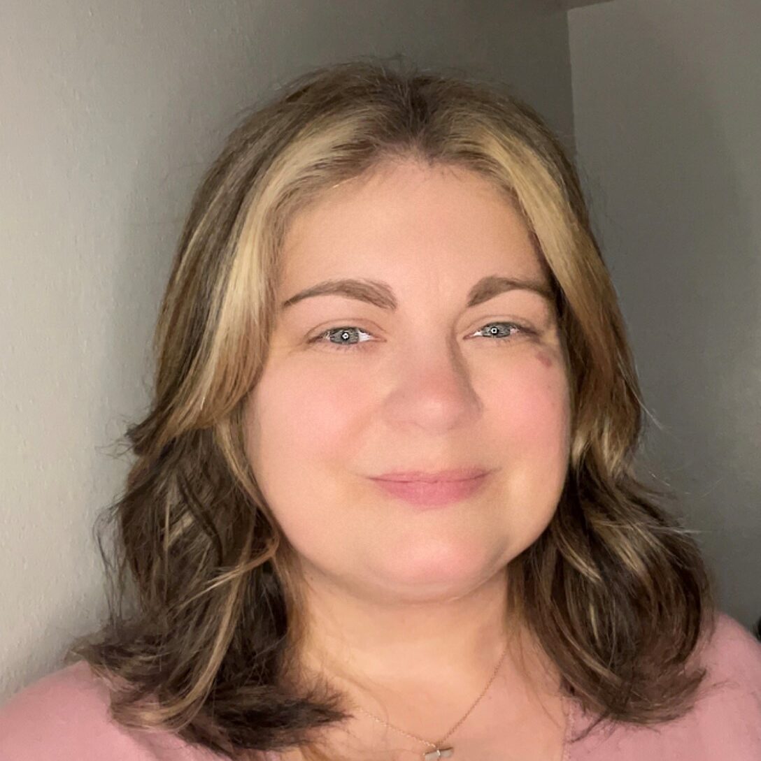 Jen is a white woman with brown shoulder length straight hair that has blonde highlights in her bangs. She is wearing a pink v neck blouse and is standing in front of a white wall.