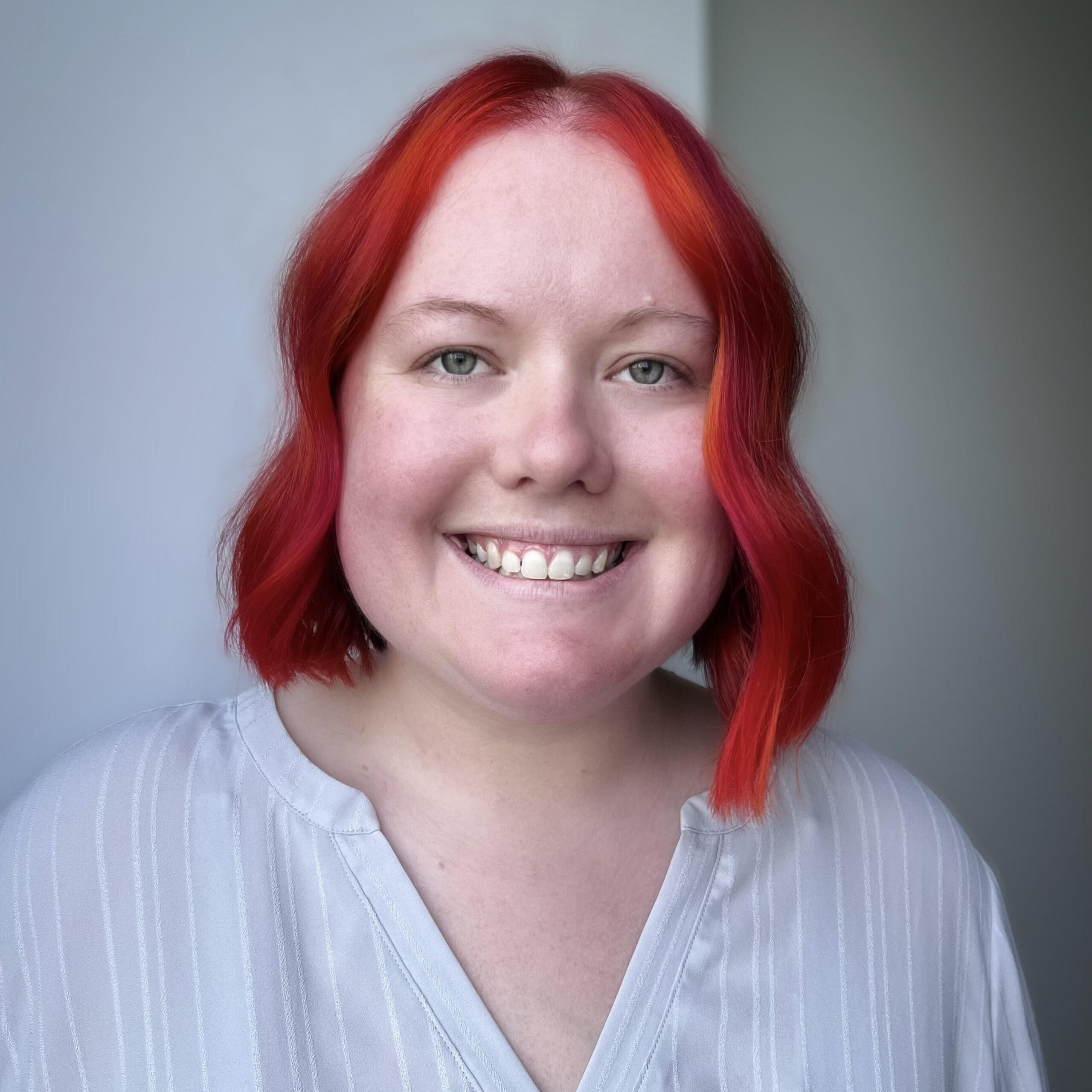 Jenny is a white woman in her late twenties with bright red hair that is cut right above her shoulders. It is curled slightly. She has bright eyes and a bright smile. She is pictured in front of a light white wall and is wearing a light white shirt.