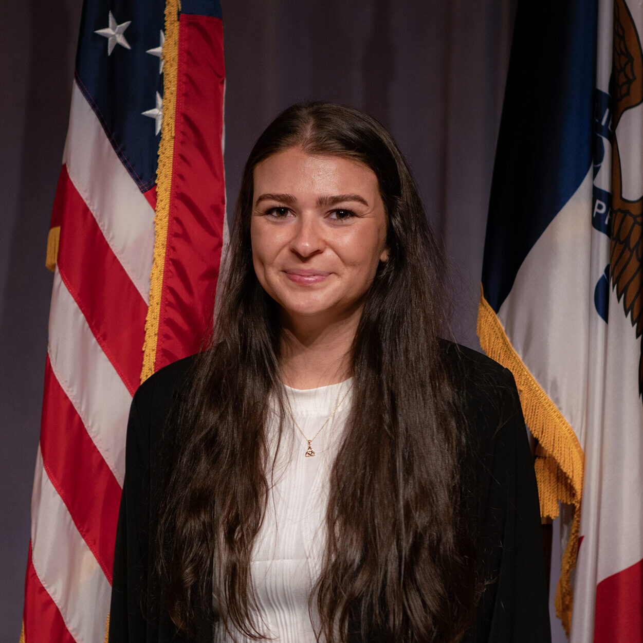 white woman with long brown hair split down the middle stands in front of an American flag, the Iowa flag and a curtained off wall.