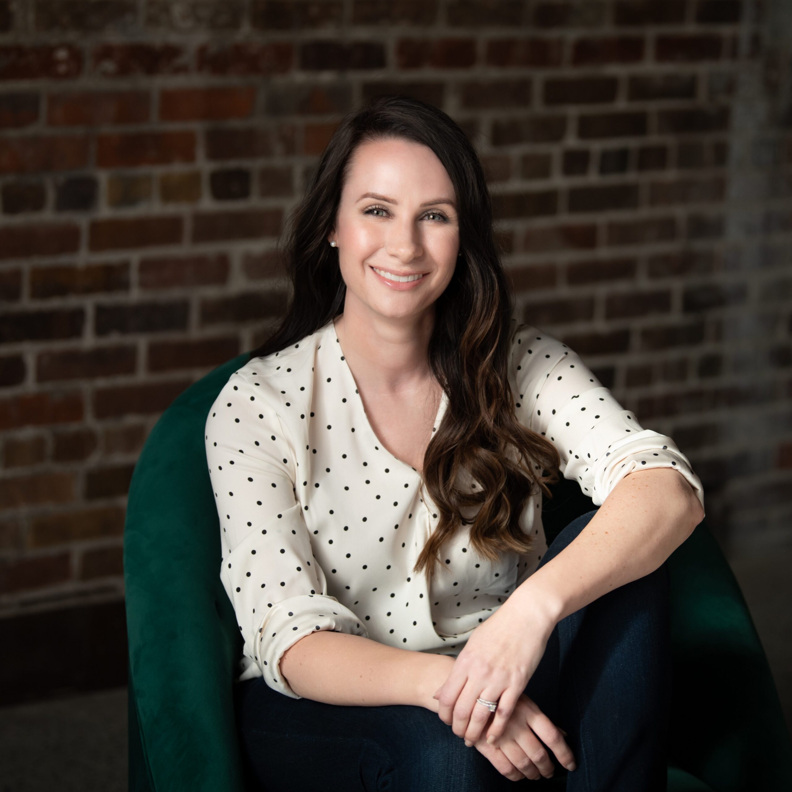 a white woman sits on a green chair in front of a brick wall. She is resting her arm on her knee wearing a white blouse. Her dark brown hair is curled and very long. She is smiling kindly at the camera